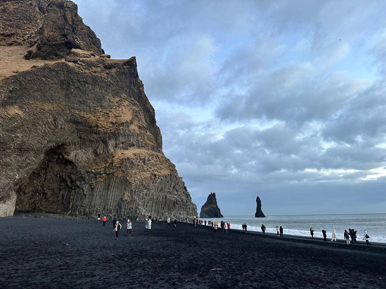 Tourists walk on the black sand beach of Reynisfjara lined by basalt columns on one side and the Reynisdrangar Sea Stacks visible in the distance.