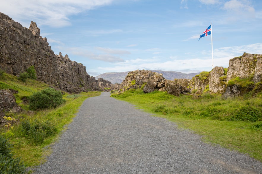 Walking path at Thingvellir National Park with Almannagja Gorge Cliffs and Icelandic flag on a sunny day.