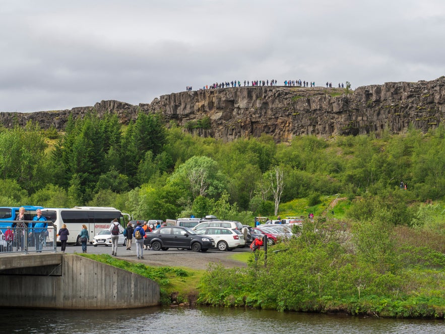 Tourists at Thingvellir National Park, Iceland, with Almannagja Cliffs, parking area, buses, and walking paths.