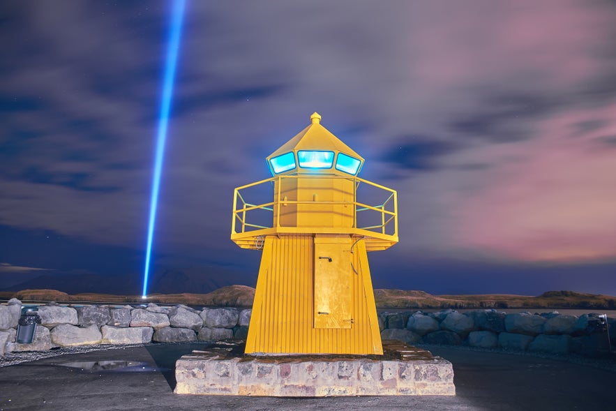 Yellow Hofdii Lighthouse illuminated at night with the Imagine Peace Tower's beam of light in the background.