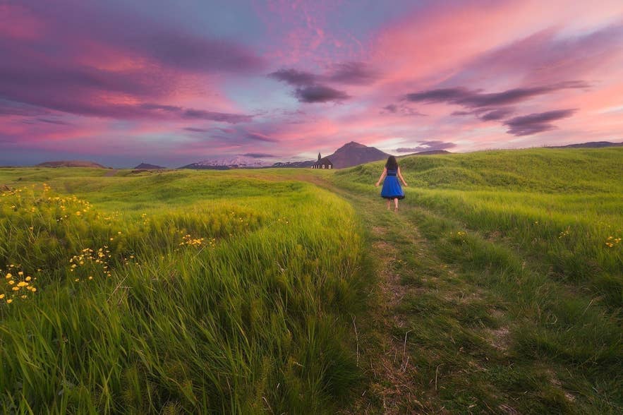 A girl wearing a blue dress walks through green fields at sunset near Budir Church in Snaefellsnes Peninsula, Iceland.