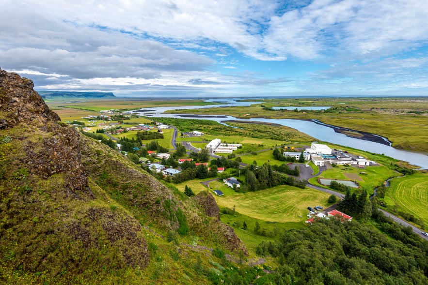 Scenic view over Kirkjubaejarklaustur village in South Iceland with a winding river, green fields, scattered houses, and distant mountains under a partly cloudy sky.