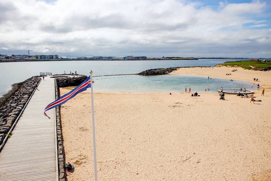 Zandstrand van het geothermische strand Nautholsvik in Reykjavik met zwemmers en een promenade langs de zee.