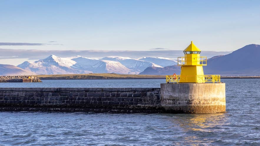 Bright yellow lighthouse in Reykjavik on coastal pier with snow-capped mountains in the background.