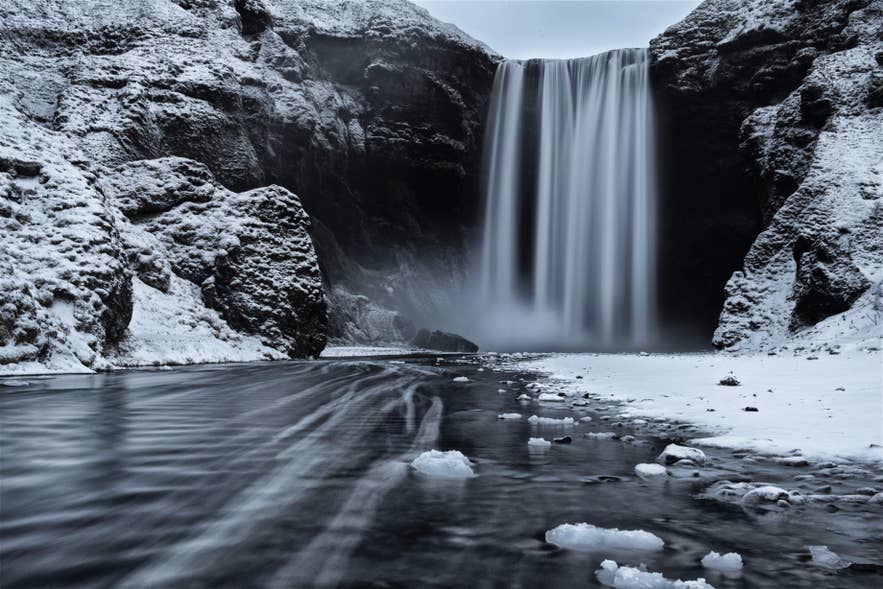 Snow-covered Skogafoss Waterfall in the South Coast flowing into an icy river in a winter landscape.