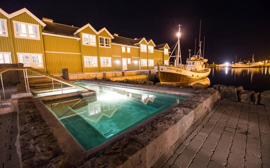 Piscina termal exterior junto al puerto de Siglo Hotel, en Siglufjörður, iluminada por la noche junto a barcos pesqueros tradicionales.