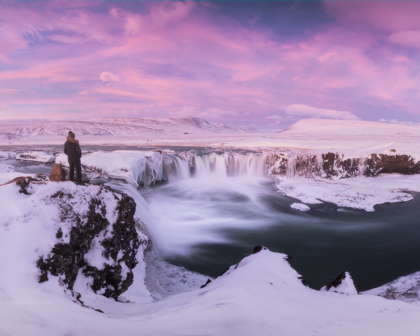 Winter view of Godafoss Waterfall under a pink sky, with a traveler standing on a snowy cliff edge.