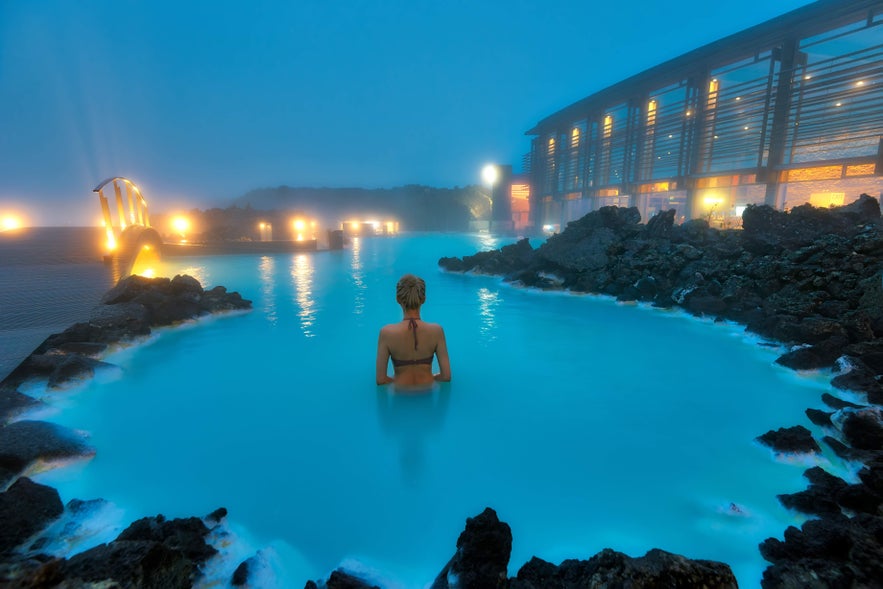 A woman in a bathing suit, with her back to the camera, relaxes in the warm, milky blue waters of the Blue Lagoon in Iceland.