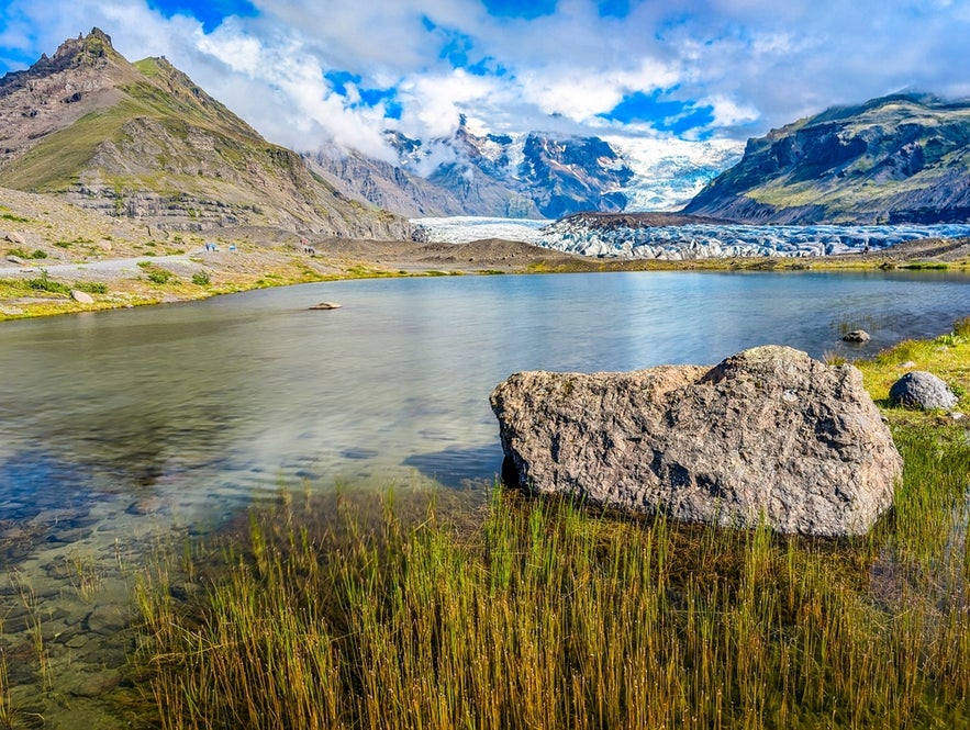 An aerial drone view shows the Svinafellsjokull glacier in Vatnajokull National Park, Iceland, surrounded by mountains and grass on a bight sunny day.