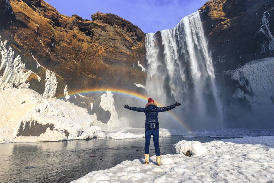 Une femme se tient devant la cascade de Skogafoss en Islande, les bras levés, tandis qu’un arc-en-ciel apparaît dans la brume de l’eau qui tombe en cascade.