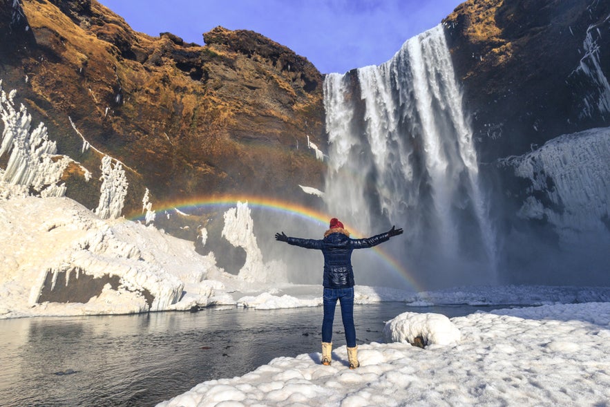 A woman stands in front of Skogafoss waterfall in Iceland with her arms raised, while a rainbow appears in the mist of the cascading water.
