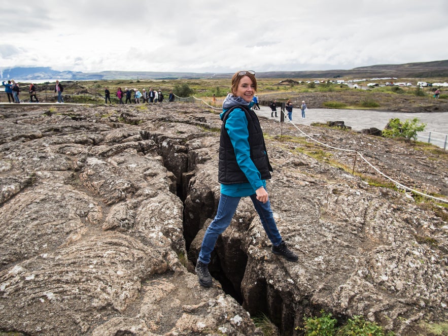 An attractive woman stands with one foot on each side of a narrow earth gap at Thingvellir National Park, a key stop on the Golden Circle route, during a weekend in Iceland.