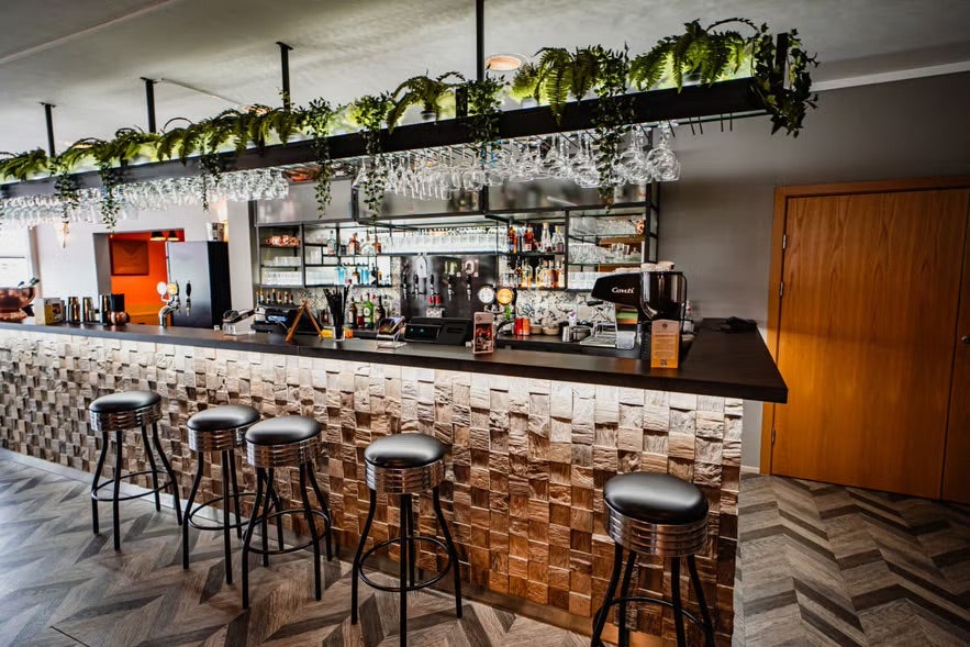 Modern bar interior at Glod Restaurant in Egilsstadir, featuring a patterned wooden bar front, black stools, hanging glasses, and greenery above the counter.