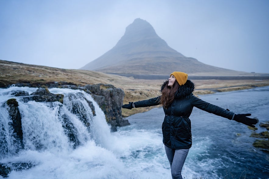 A happy, carefree woman dances with open arms in the winter wind during a weekend in Iceland, surrounded by nature, with Kirkjufell Mountain in the background.