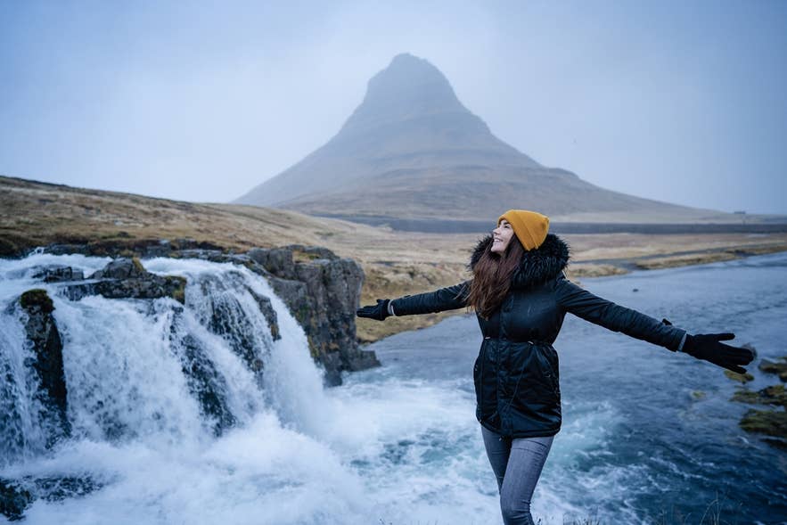 Szczesliwa, beztroska kobieta tanczy z szeroko rozlozonymi ramionami na zimowym wietrze podczas weekendu na Islandii, otoczona natura, z gora Kirkjufell w tle.