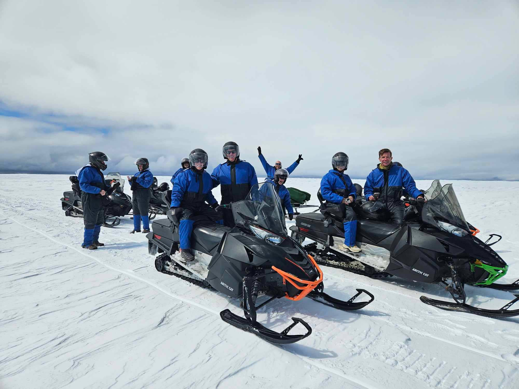 A group in blue suits prepares for a Langjokull Glacier snowmobile tour under bright midday skies.