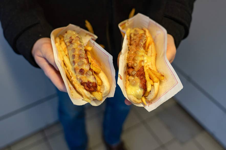 Two Icelandic hot dogs topped with fries, onions, and melted cheese, freshly prepared and held by a person indoors