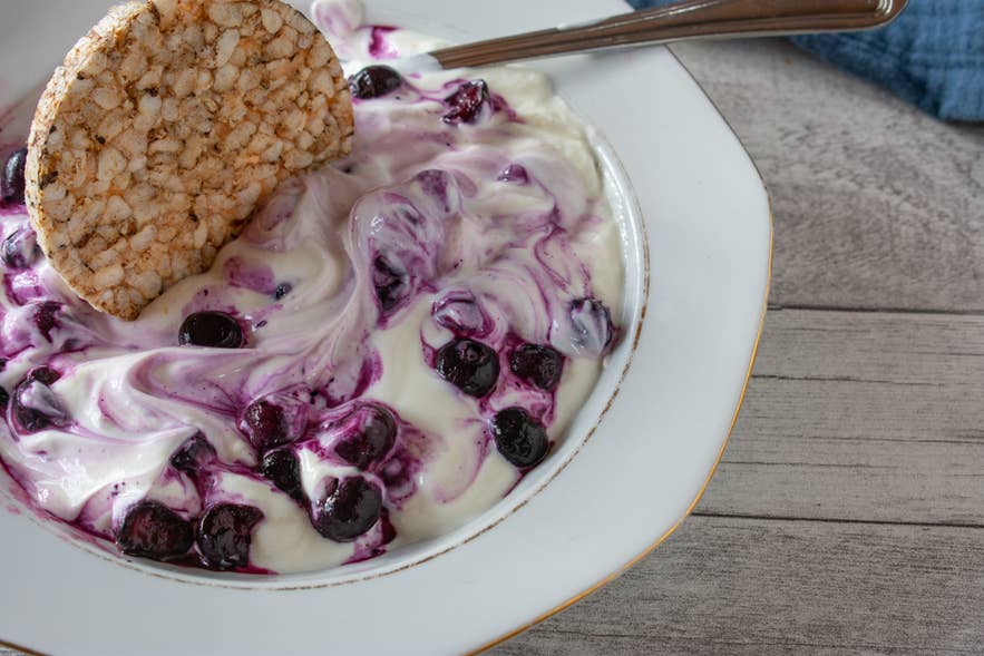 IJslandse skyr-yoghurt met blauwe bessen, geserveerd in een witte kom met een rijstwafel en lepel op een houten tafel.