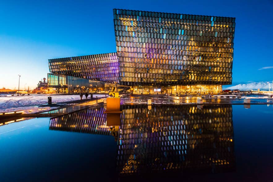 Concertgebouw Harpa in Reykjavik verlicht tijdens de schemering. De glazen gevel weerspiegelt kleurrijke lichten op het kalme water ervoor, met sneeuw en de haven op de achtergrond.