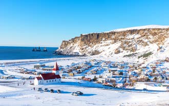 Snötäckt byn Vik i södra Island, med sin rödvita kyrka som blickar ut över färgglada hus, branta havsklippor och Reynisdrangar-klipporna som reser sig ur Nordatlanten under en klarblå vinterhimmel.