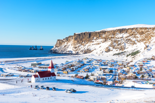 Snow-covered village of Vik in South Iceland, with its red-roofed church overlooking colorful houses, steep sea cliffs, and Reynisdrangar sea stacks rising from the North Atlantic under a clear blue winter sky.