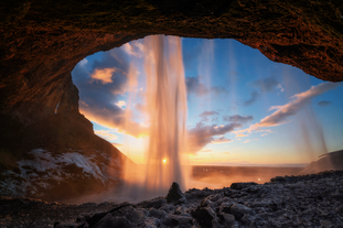 View from behind Seljalandsfoss waterfall in South Iceland at sunset, with golden light shining through the falling water and illuminating the icy rocks and winter landscape beyond.