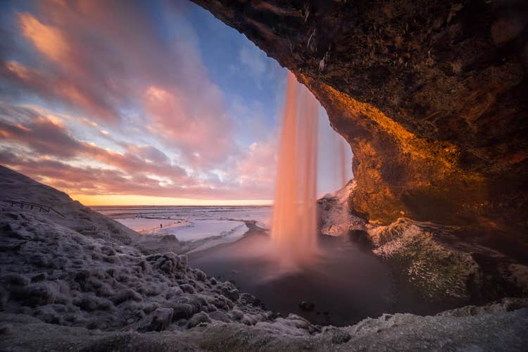 Seljalandsfoss waterfall in South Iceland viewed from behind the cascade at sunset, with warm light illuminating the icy rocks and snow-covered landscape beneath a colorful winter sky.