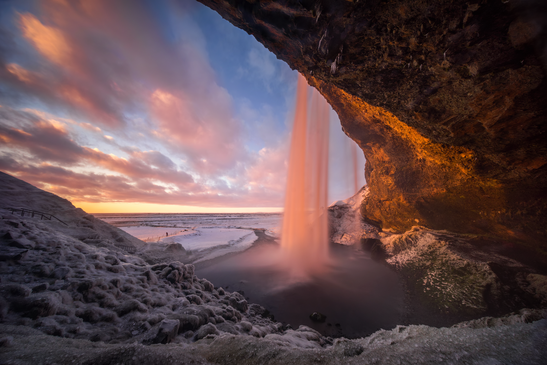 La cascada Seljalandsfoss, en el sur de Islandia, vista desde detrás de la cascada al atardecer, con una cálida luz que ilumina las rocas heladas y el paisaje cubierto de nieve bajo un colorido cielo invernal.