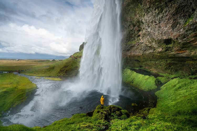 A person in a yellow jacket stands behind Seljalandsfoss Waterfall surrounded by green cliffs.