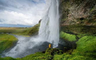 Une personne vêtue d’une veste jaune se tient derrière la cascade de Seljalandsfoss, entourée de falaises verdoyantes.
