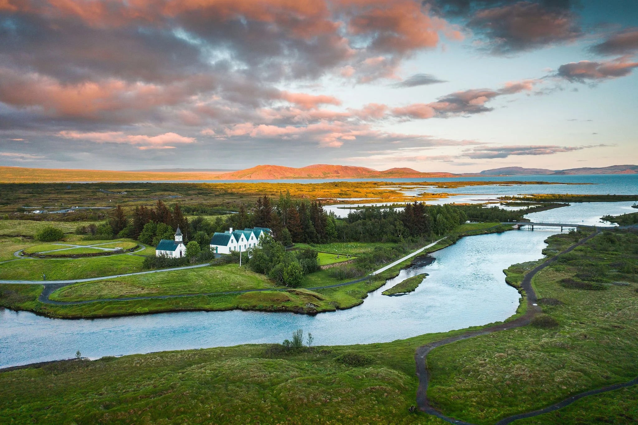 Aerial view of Thingvellir National Park with rivers, green fields, and historic white buildings under colorful skies.