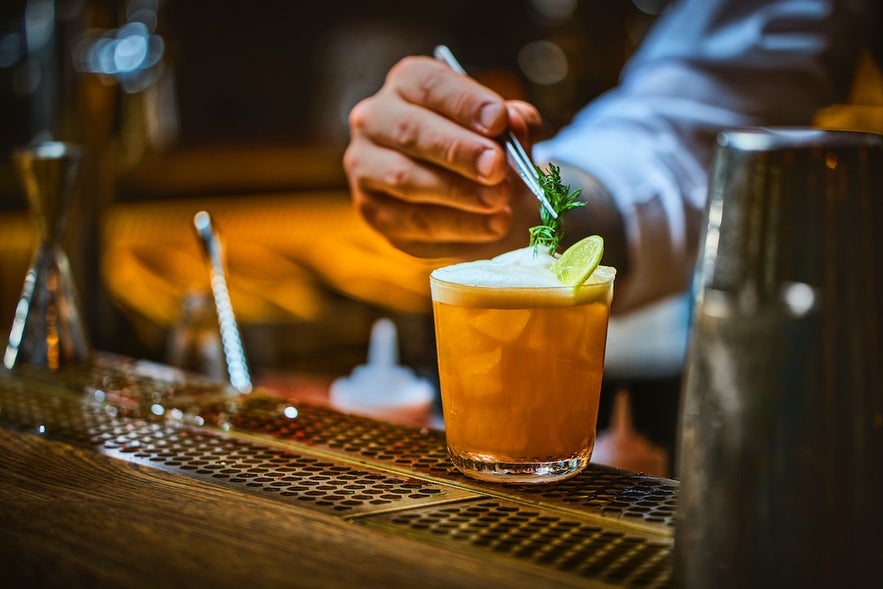 Bartender at Tipsy Bar & Lounge in Reykjavík garnishes an orange cocktail with rosemary and lime on a metal counter.