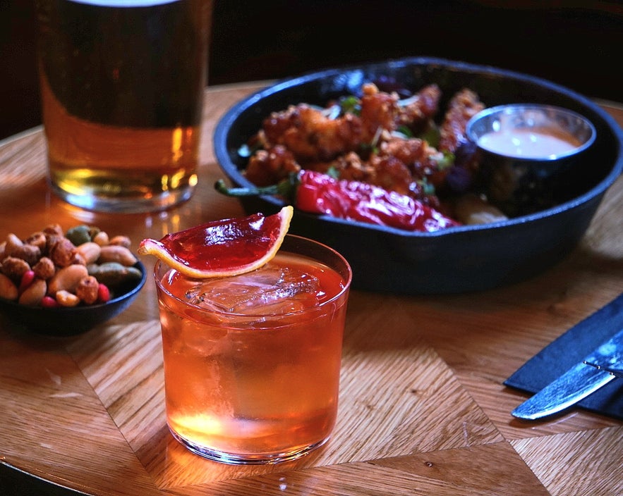 Cocktail with blood orange slice, fried chicken, nuts, and beer on a wooden table at Tipsy Bar & Lounge in Reykjavík, Iceland.