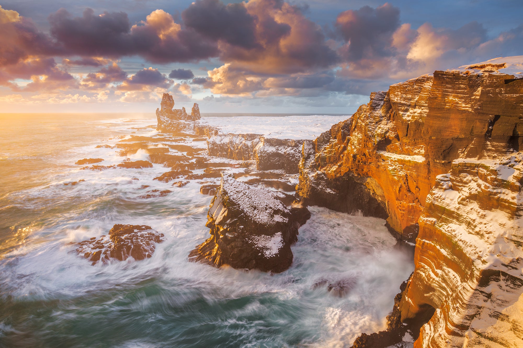 Londrangar Cliffs glowing at sunset as waves crash against snow-covered volcanic rock on Snaefellsnes Peninsula.