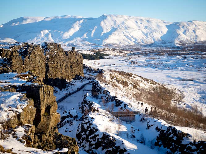 Snowy cliffs and hikers exploring Thingvellir National Park with distant mountains glowing under clear winter sunlight.