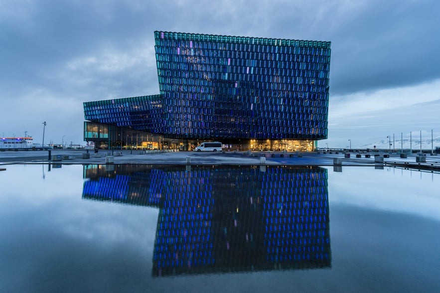 Harpa Concert Hall in Reykjavik reflects on calm water at dusk, its glass facade glowing blue against the Icelandic sky.