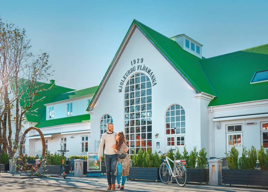 A couple walks past the white building with a green roof of Mjolkurbu Floamanna in Selfoss.