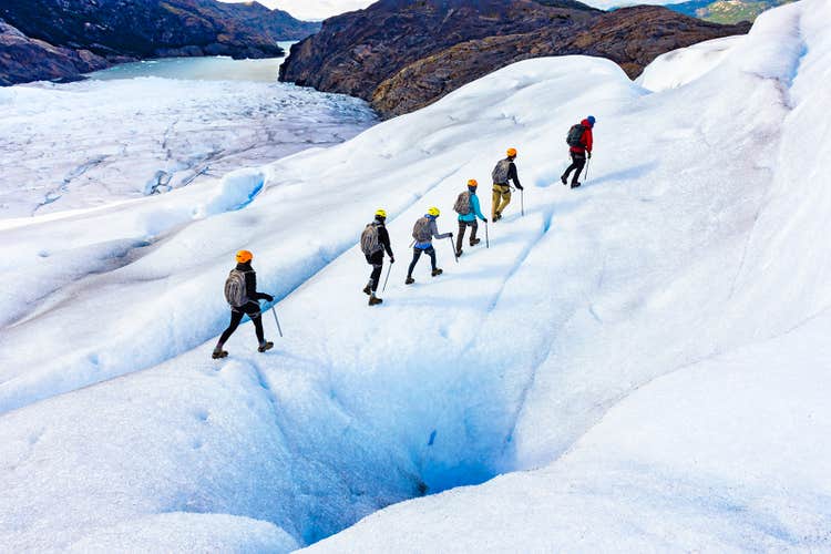 Group of hikers with helmets and ice axes trekking across Skaftafell Glacier with a frozen lake and cliffs below.
