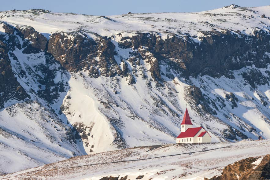 Die kleine weiße Kirche Vikurkirkja mit rotem Dach steht auf einem verschneiten Hügel vor schroffen Bergen im Süden Islands.