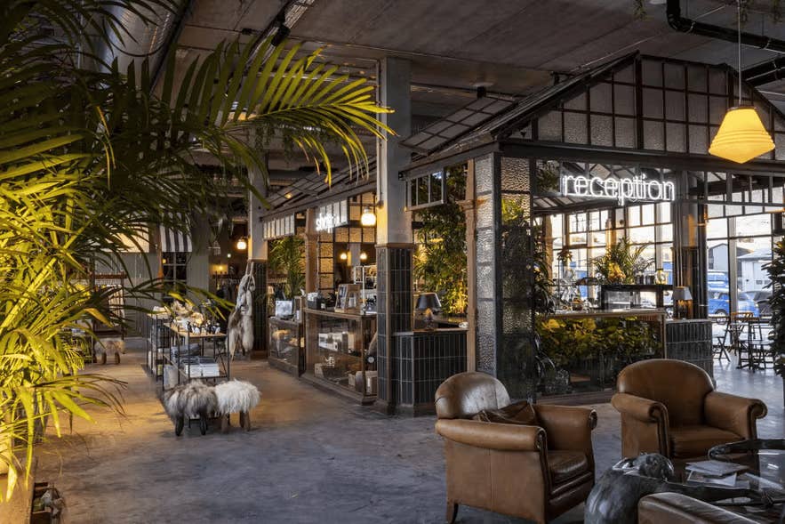 Hotellobby mit warmem Licht, Ledersesseln, üppigen Pflanzen und einer stylischen Rezeption mit Neon-Schriftzug und modernem Industriedesign.
