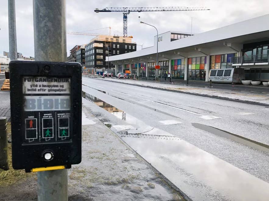 Regennasse Straßenszene vor dem Flohmarkt Kolaportid in Reykjavik, Island – im Vordergrund ein Fußgängerampelknopf, dahinter Kräne und moderne Gebäude.