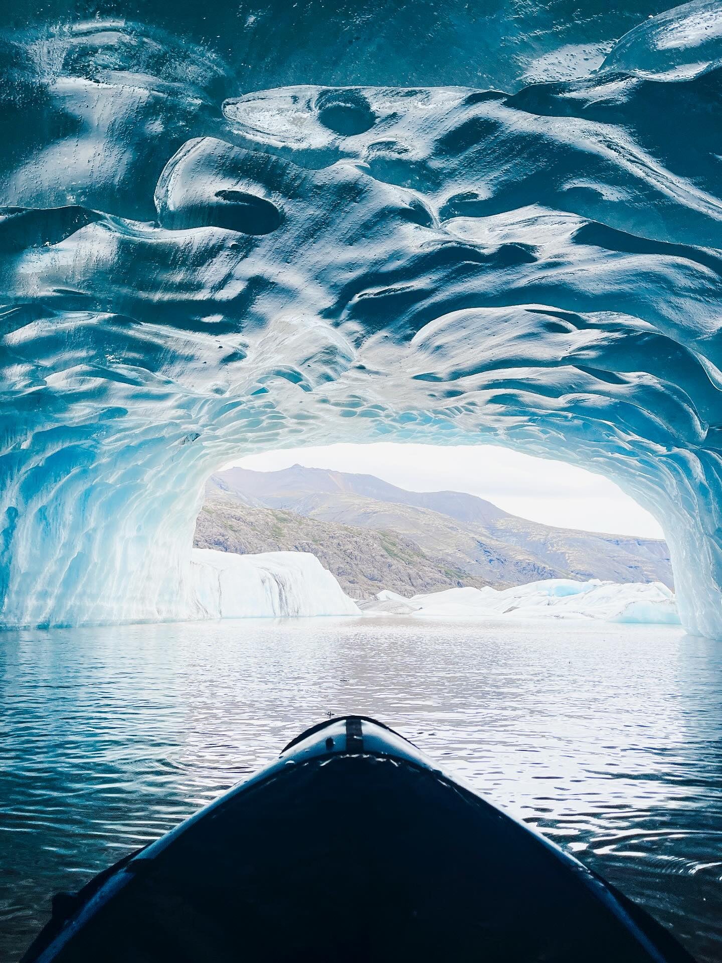 Kayaking inside a blue ice cave in Iceland with glacier lagoon views and rugged mountain landscape beyond.