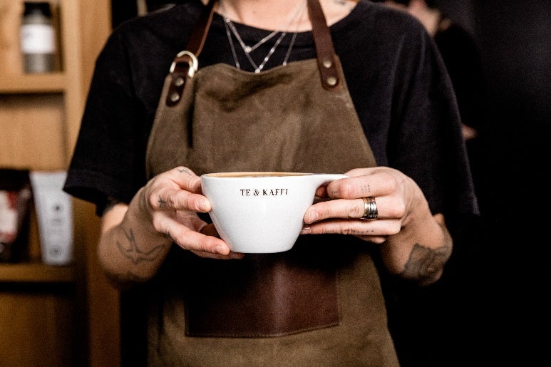 A barista wearing a brown apron holds a white coffee cup labeled "TE & KAFFI" in both hands.