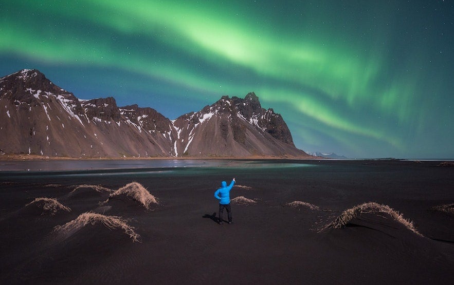 Person watching northern lights over Vestrahorn mountain in Iceland, with green aurora above black sand beach in September