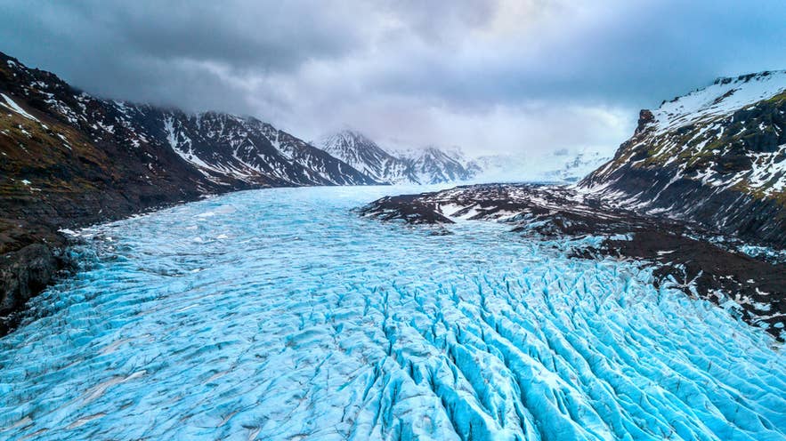Vista aerea del ghiacciaio Vatnajokull nel sudest dell’Islanda, con ghiaccio blu brillante e montagne innevate sotto cieli nuvolosi.