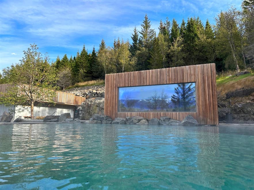 Геотермальный бассейн Forest Lagoon в Исландии: над водой поднимается пар, рядом расположены современный спа-комплекс и покрытый лесом склон под ясным небом.
