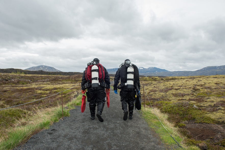 Two scuba divers walking toward Silfra Fissure in Thingvellir National Park, Iceland, carrying diving gear.