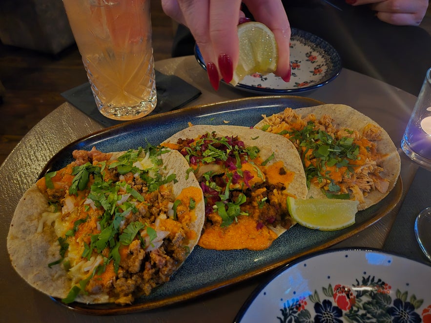 Close-up of three tacos topped with herbs and sauce, with a hand squeezing lime juice and a cocktail beside the plate.