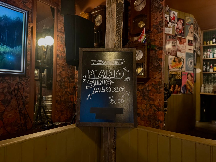 Chalkboard sign inside Elly Bar advertising a piano sing-along night, surrounded by vintage posters and warm lighting.
