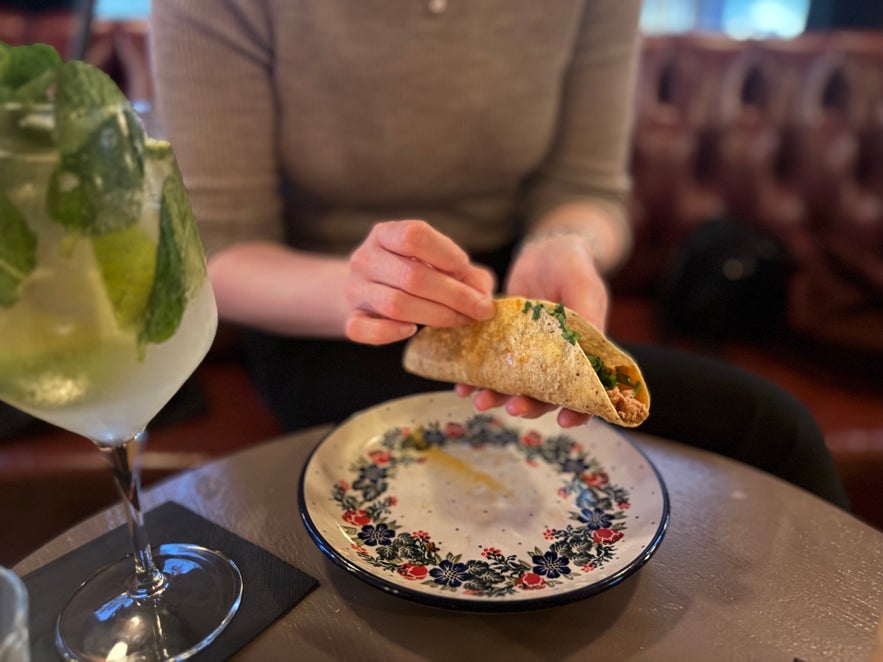Person holding a taco above a decorative plate at Elly Bar, with a mint cocktail on the table.